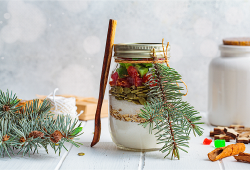 A mason jar filled with baking ingredients, layered and topped with dried fruit and seeds, decorated with a pine branch and cinnamon stick. A spool of twine, pine sprigs, and a jar are on the white wo