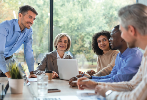 A group of five diverse professionals are gathered around a table in a bright office, engaged in discussion. One man stands, while the others sit with laptops, papers, and coffee, all appearing attent