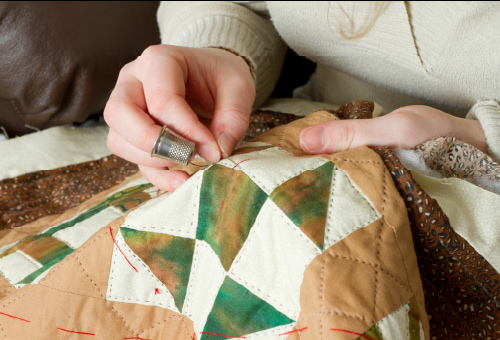 A person wearing a thimble sews stitches on a colorful patchwork quilt, focusing on the detailed needlework with both hands.